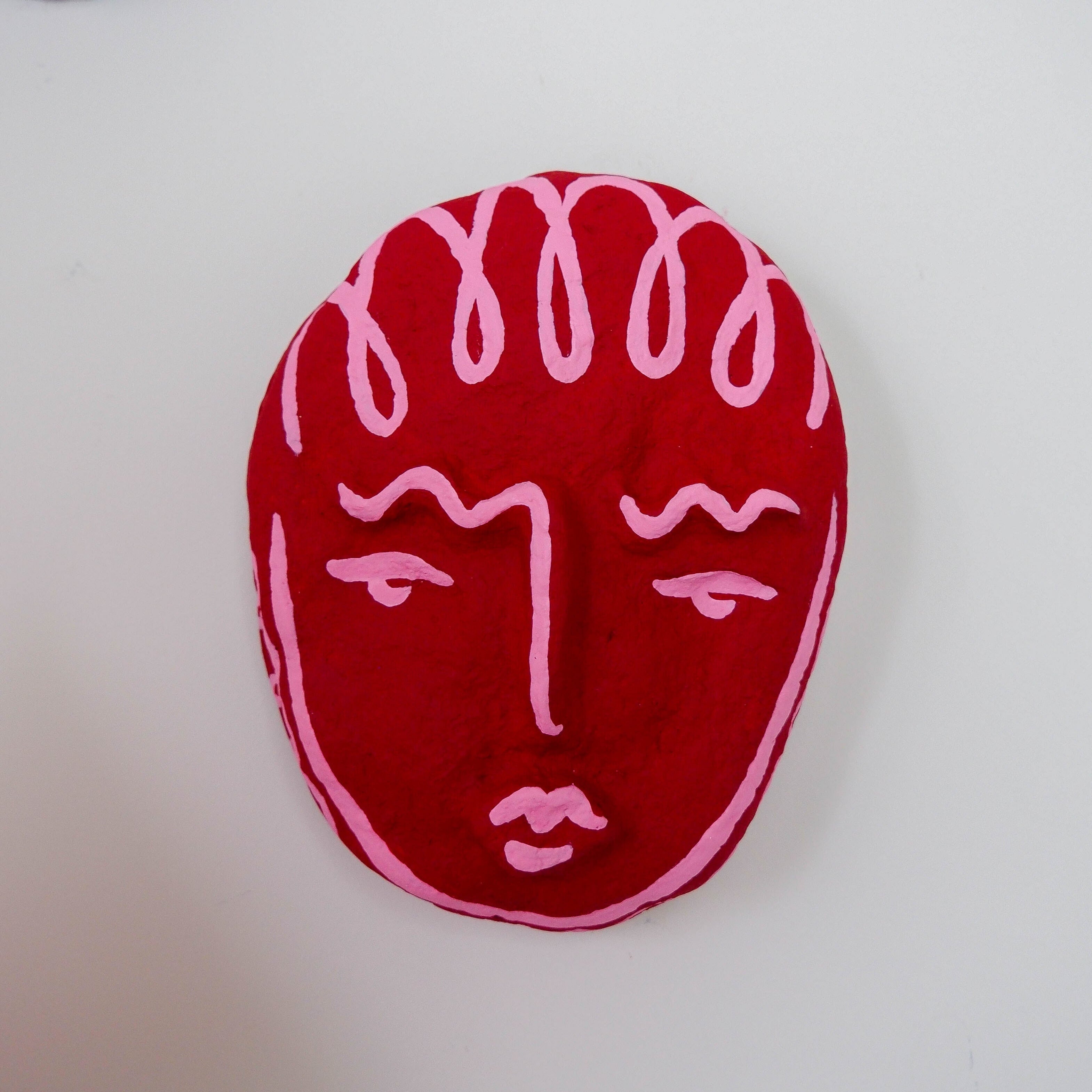 Red face-shaped mask sculpture with pink lines on a white background